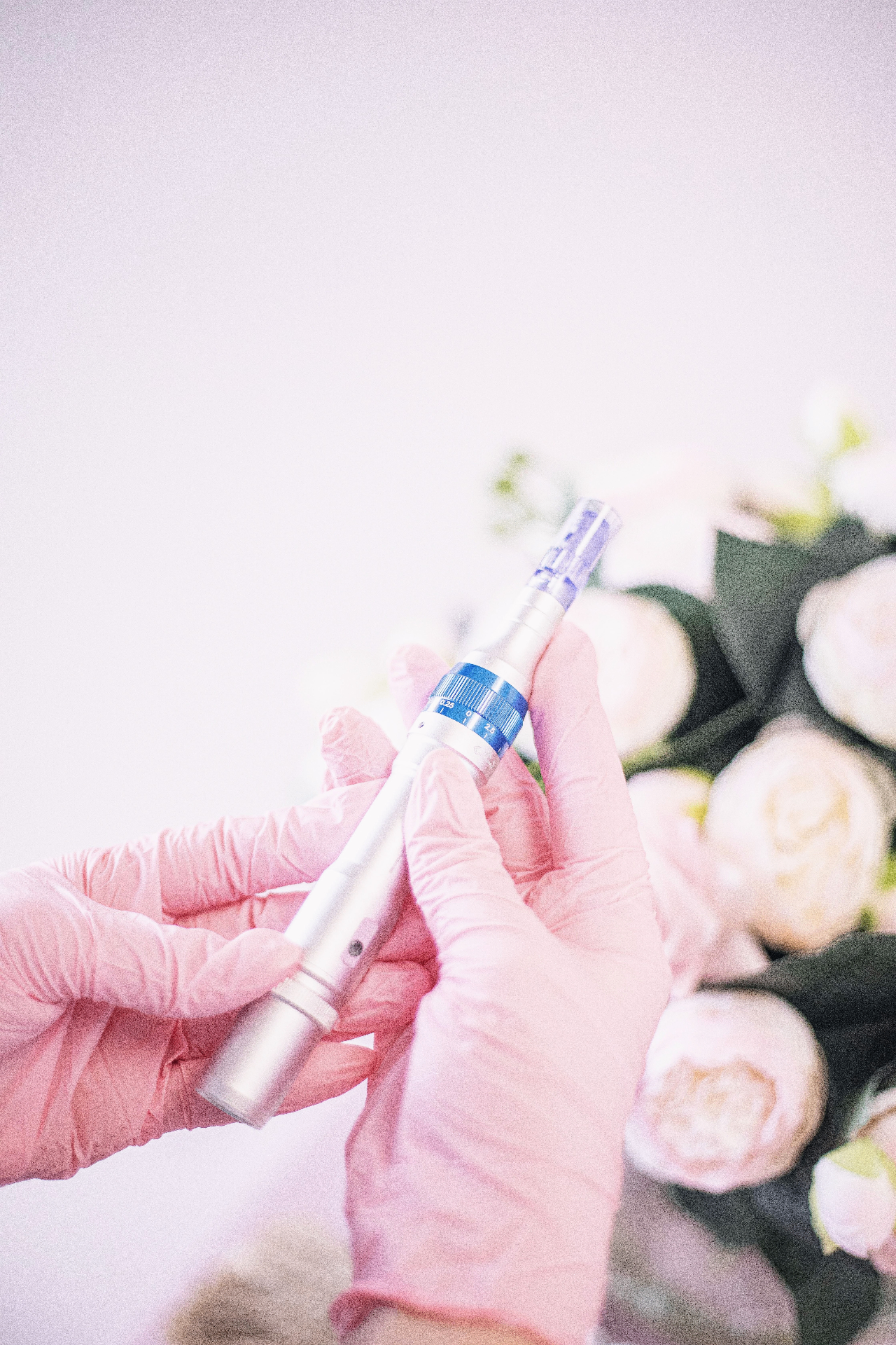 Gloved hands holding a microneedling pen beside soft pink roses
